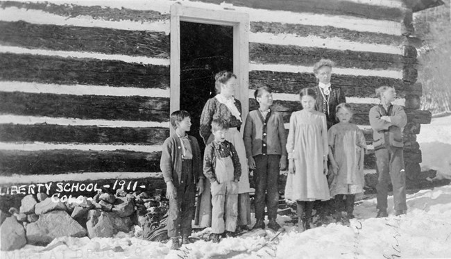 A log schoolhouse with two teachers and students standing in front