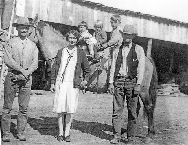 Historical black and white photo of adults and children on a ranch. The children are sitting on horseback.