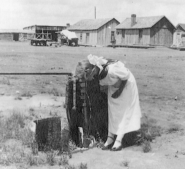 Historical black and white photo of a girl in formal white attire drinking from a water spigot near ranch buildings