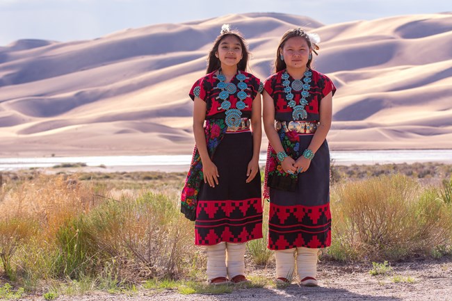 Two young Dineh Tah' Navajo Dancers in colorful regalia stand in front of the dunes