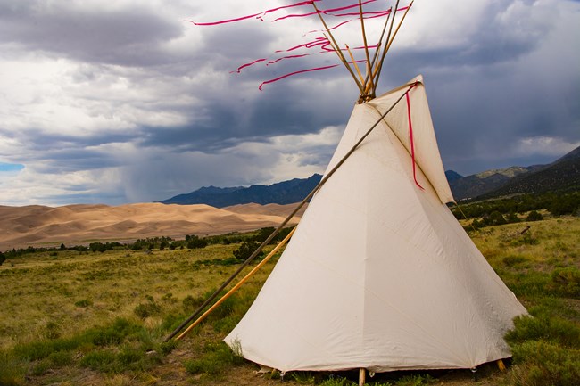 A tipi stands in front of the dunes and mountains beyond