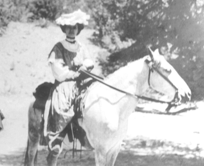 Historical black and white photo of Julia Herard, a young woman in dress attire riding on a horse near the dunes