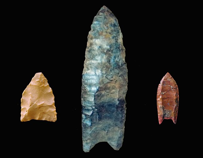 Three paleo projectile points are displayed on a black background. On the left is a small, triangular Pre-Clovis point, in the center is a long Clovis spearpoint; on the right is a small fluted Folsom point