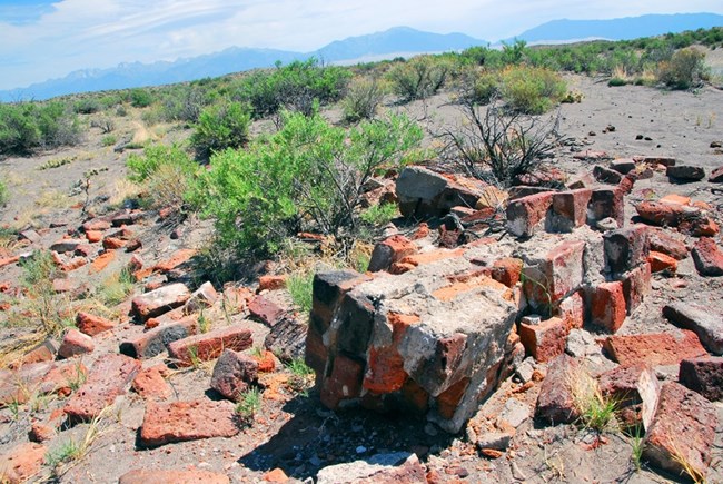Amid desert shrubs and sand, broken sections of brick wall are strewn around. In the distance are dunes and mountains.