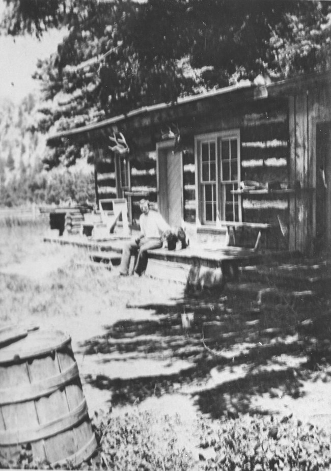 Historical black and white photo of a large log cabin with Ulysses Herard sitting in front