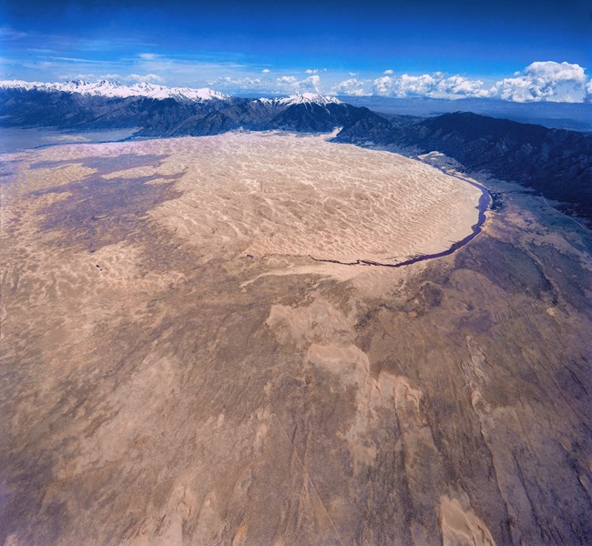 An aerial view of the dunefield, showing grasslands, small dunes migrating toward the dunefield, creeks on the sides, and snowy mountains