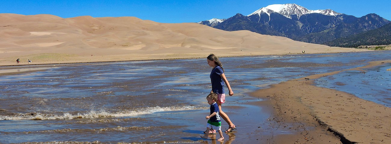 A teen girl and her toddler brother walk along the edge of Medano Creek during peak flow in late spring. A surge flow wave is moving in front of them, and another one is approaching farther up the creek. Above the creek are the dunes and a snowy mountain.