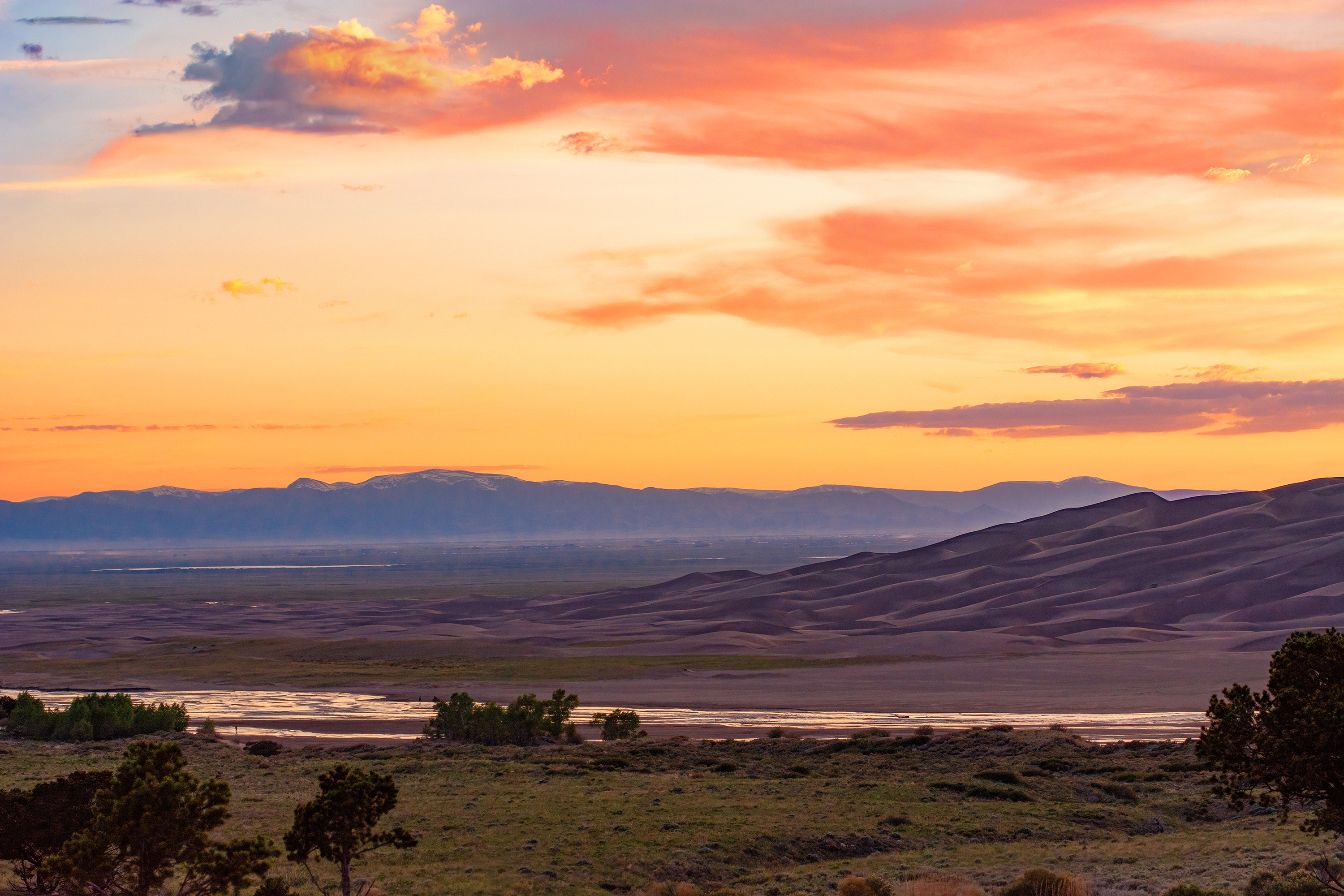 A summer view of the San Luis Valley, with the dunes surrounded by grasslands, trees, Medano Creek, and wetlands. In the distance are the rounded forms of the San Juan Mountains. An orange sunset illuminates the sky.