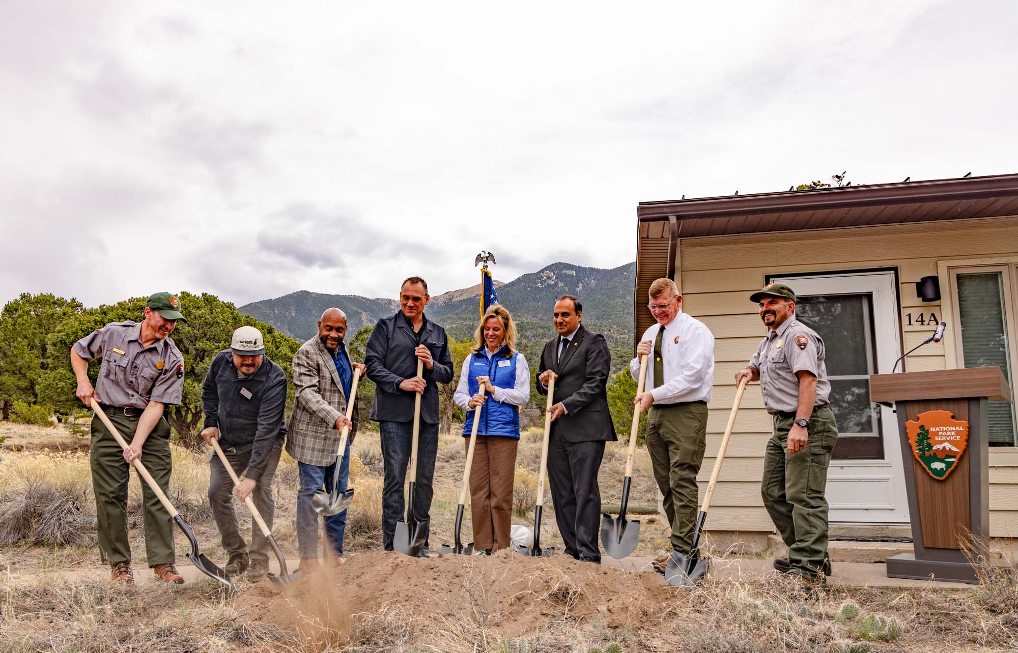 Eight people stand with shovels next to a pile of soil while out in front of a house