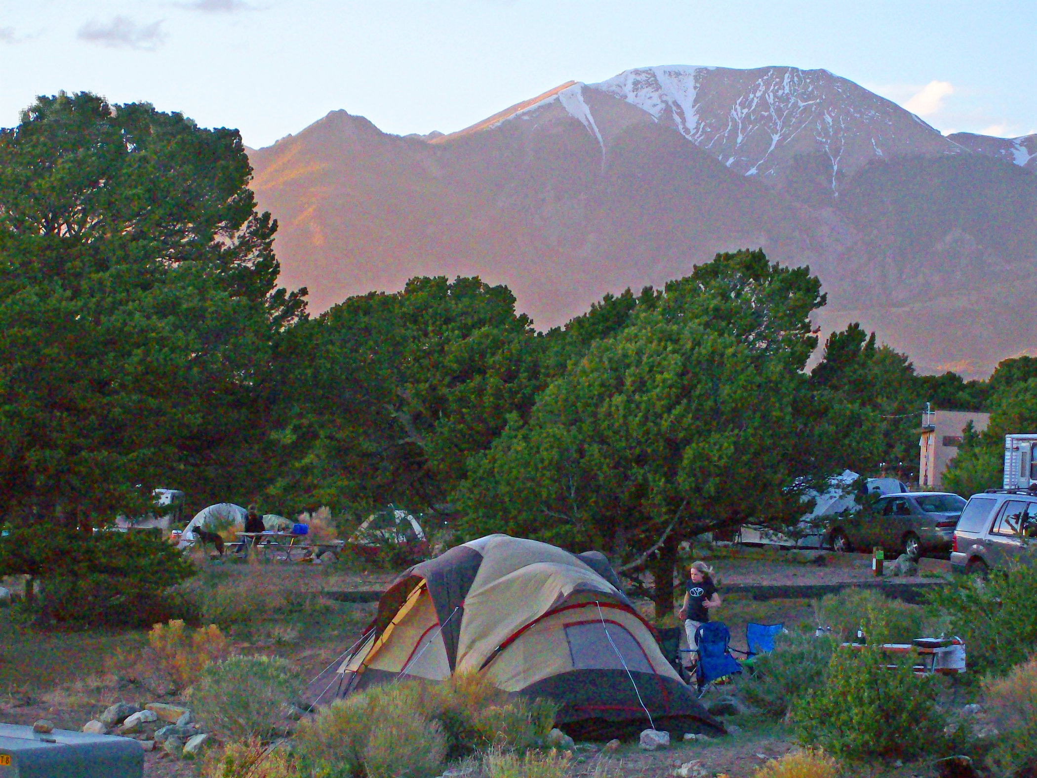 Pinon Flats Campground in Evening A girl walking to a tent with other tents, trees, and snowcapped mountain