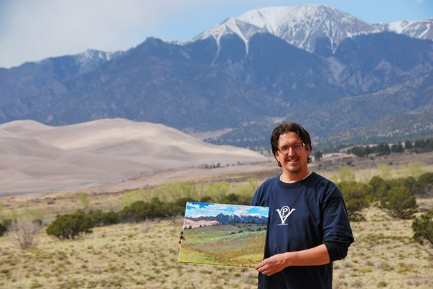 NPS photo: Artist Randy Pijoan led a painting workshop and printing demonstrations at Great Sand Dunes in 2013. In this photo taken just after his workshop, he displays a painting of the grasslands, dunes and Mt. Herard with the scene behind him.