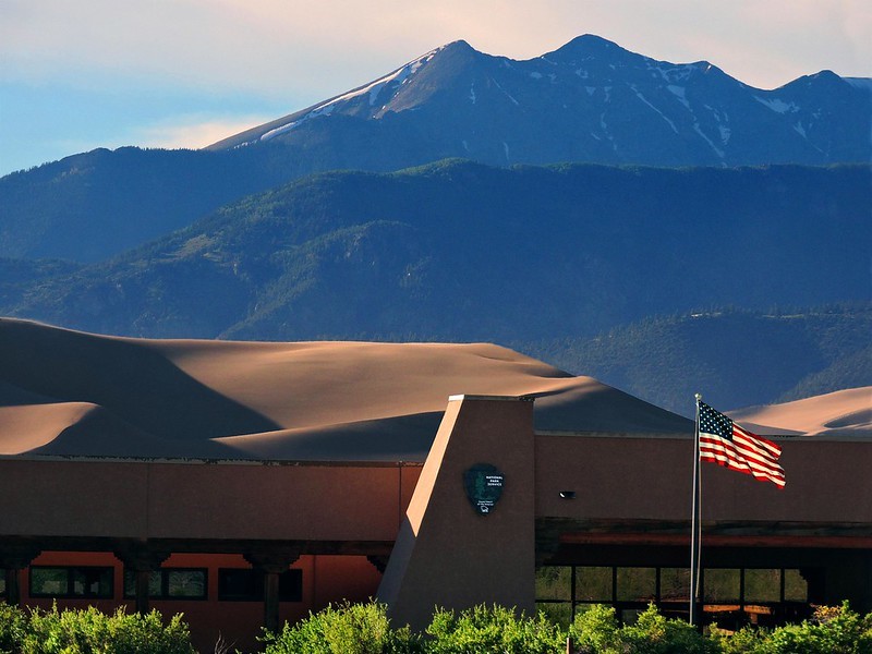 A photo of the visitor center.  The dunes and mountains rise above the building in the background.  In front of the visitor center, a row of bushes, and the U.S. flag is visible.