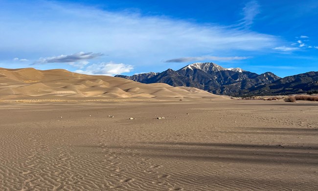 A flat sandy foreground with San Dunes and Mountains will some snow in the background under a blue sky