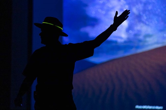 A silhouetted ranger gestures toward a large screen with an image of the dunes and night sky