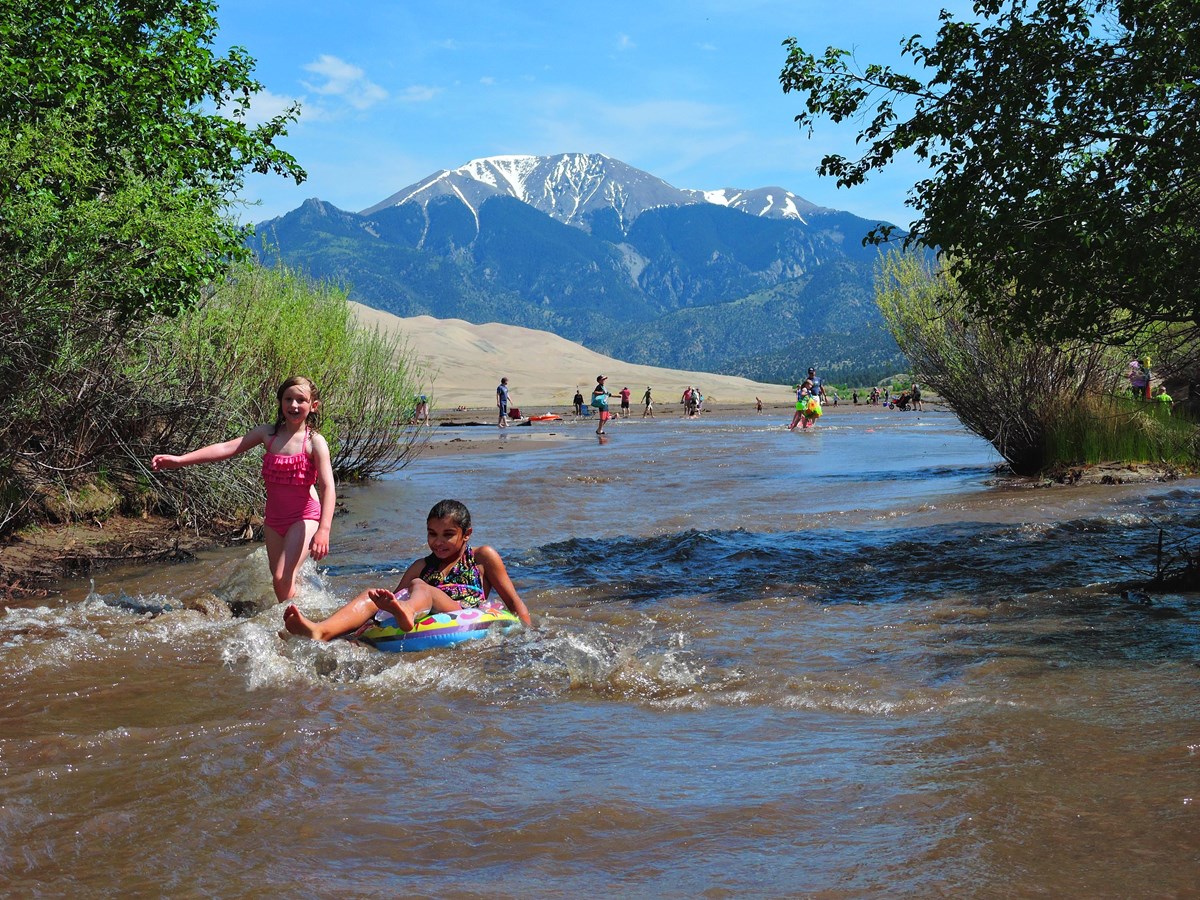 Two girls splash and float in a creek, with trees, dunes and mountains