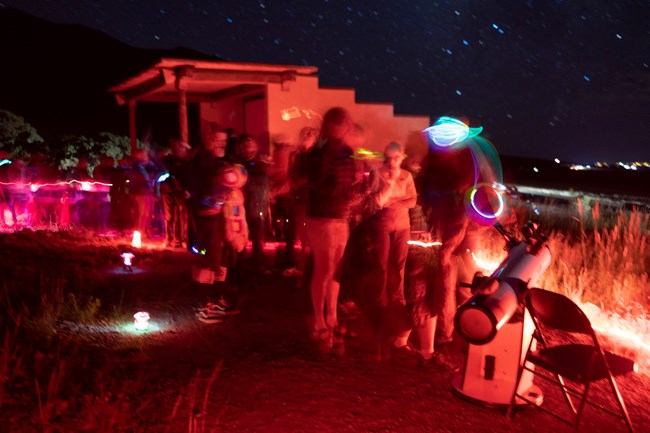A long exposure photo shows a line of visitors at the park amphitheater waiting to peer through a telescope. The scene is bathed in red light.