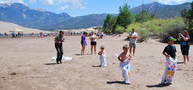 A ranger leads kids in hopping across the sand with dunes and mountains in the background
