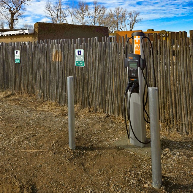 An EV charging station outside the wooden fence and adobe buildings of Fort Garland Museum