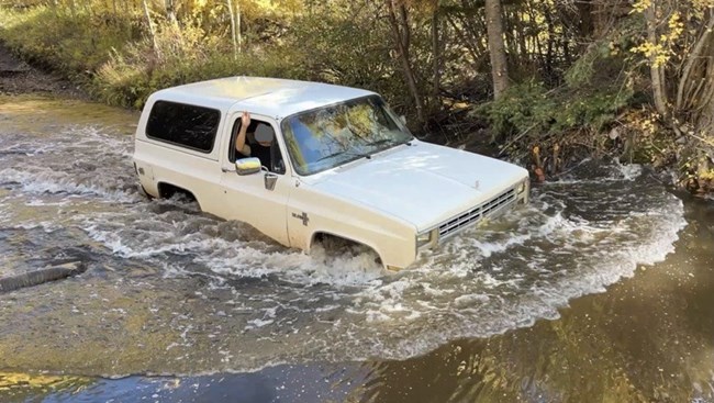 An older model SUV driving through deep water with water covering half og the front grille.