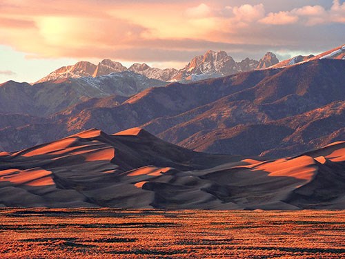 A huge dune below snow-capped mountains at sunset