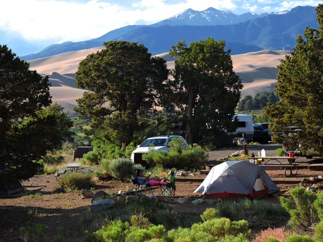 Campsite with tents and vehicles in a pinon pine forest with sand dunes and high mountains in the background