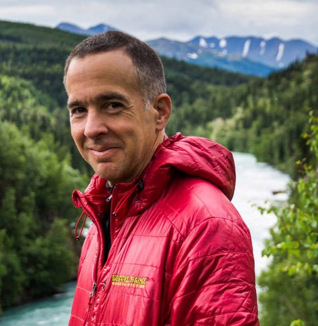 Composer Stephen Lias stands on a ridge with forest, river and a mountain behind him
