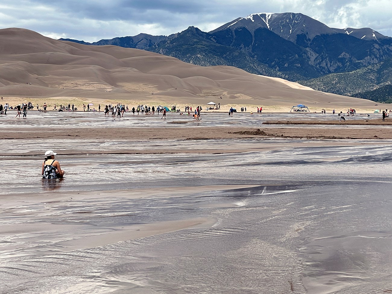 A wide shallow stream flowing at the base of dunes and mountains with visitors playing