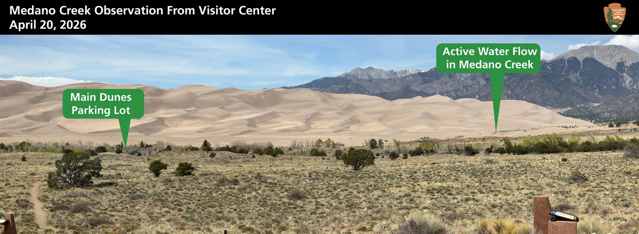 A panoramic image of sand Dunes, mountains and text labeling where dunes parking lot is and where the end of the Medano Creek is