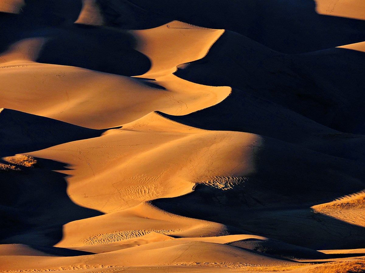Curving forms of dunes at sunset, with tiny hikers visible near the top