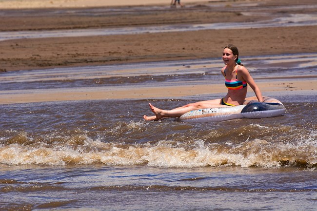 A teen girl floats on an inflatable tube down a creek with a wave flowing across sand