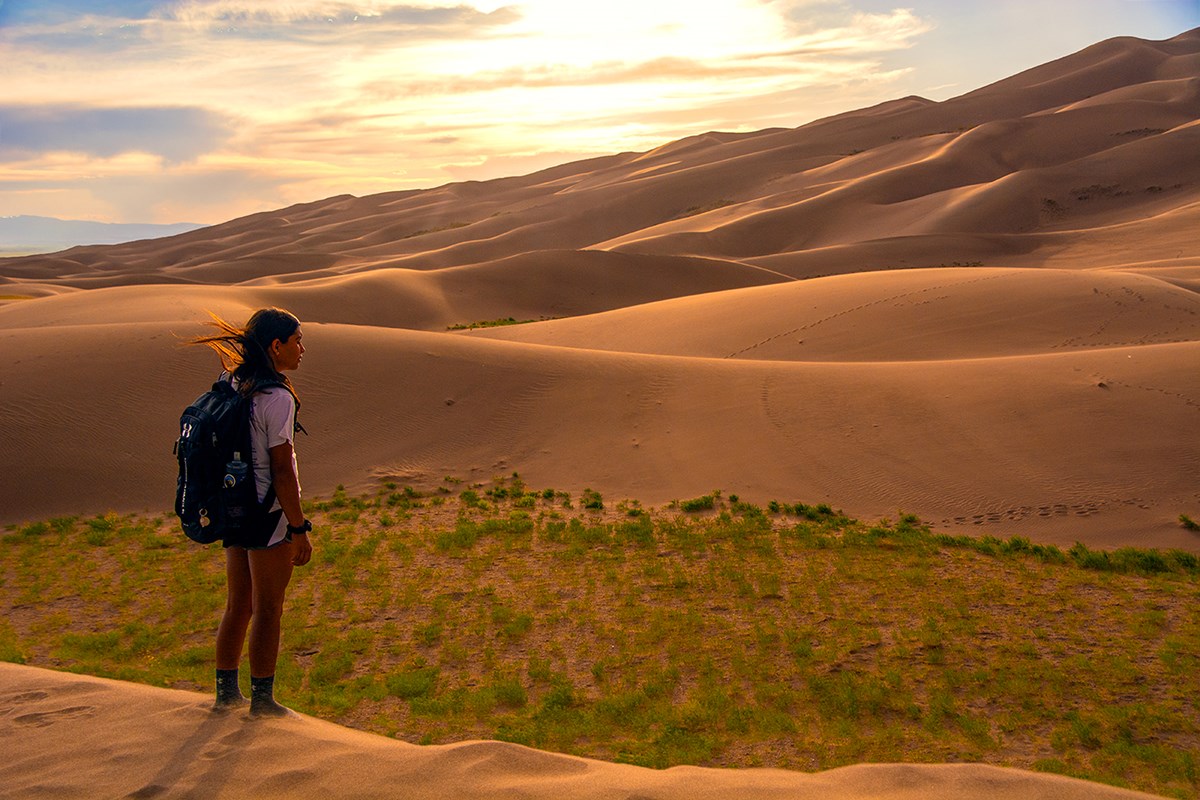 A girl with a daypack stands on the dunes with sunset light touching ridges of sand beyond her
