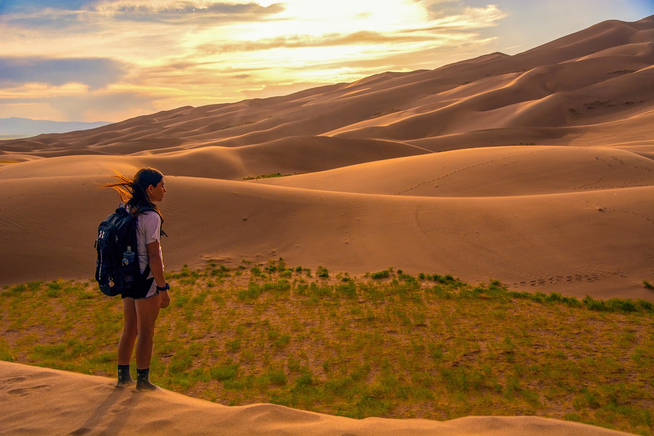 A teen girl with a backpack stands on the dunes at sunset