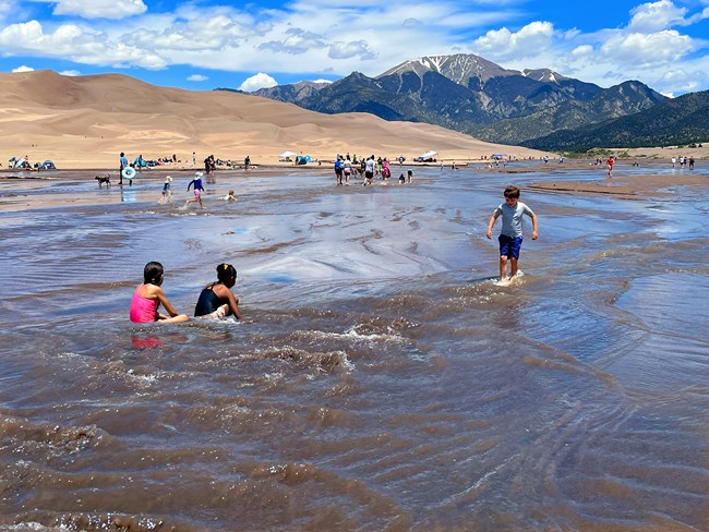 A wide creek with small waves, visitors playing in the water, dunes and snow-capped mountain in the background