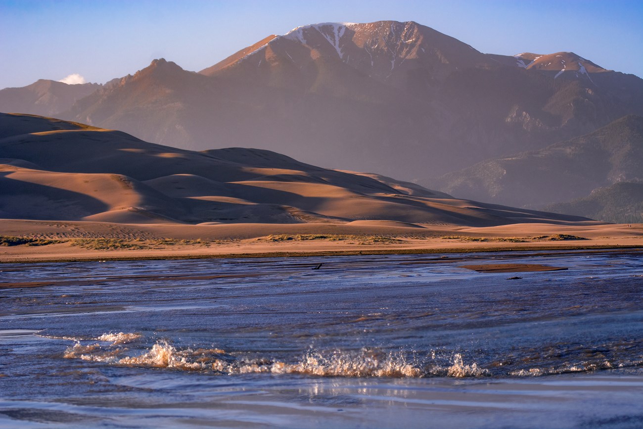 A wide shallow stream with a whitecap wave flowing at the base of dunes and mountain