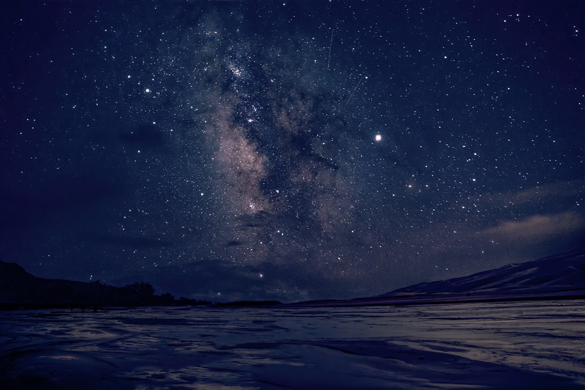 The Milky Way and other stars glow over a wide, shallow stream, with silhouetted trees at left and dunes on the right.