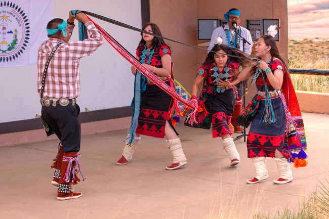 Three female Navajo dancers in colorful regalia weave a large sash while two Navajo men hold the sash and sing