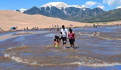 Medano Creek with waves, people playing in water, dunes, and snowcapped mountain