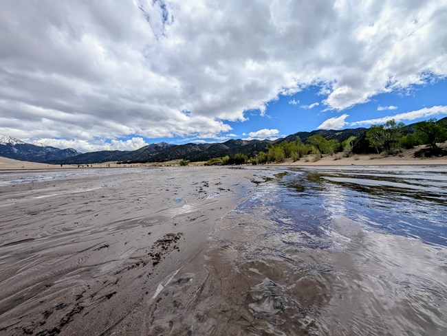 A shallow creek courses across a flat sandy area with people, trees, dunes and mountains in the background