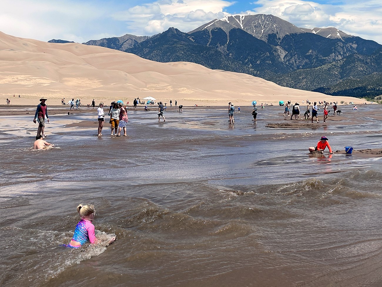A wide shallow stream flowing at the base of dunes and mountain with lots of visitors playing