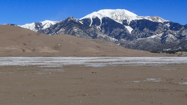 A few of a dry sandy creek bed with partial snow cover, with dunes and a snow-covered mountain in the background