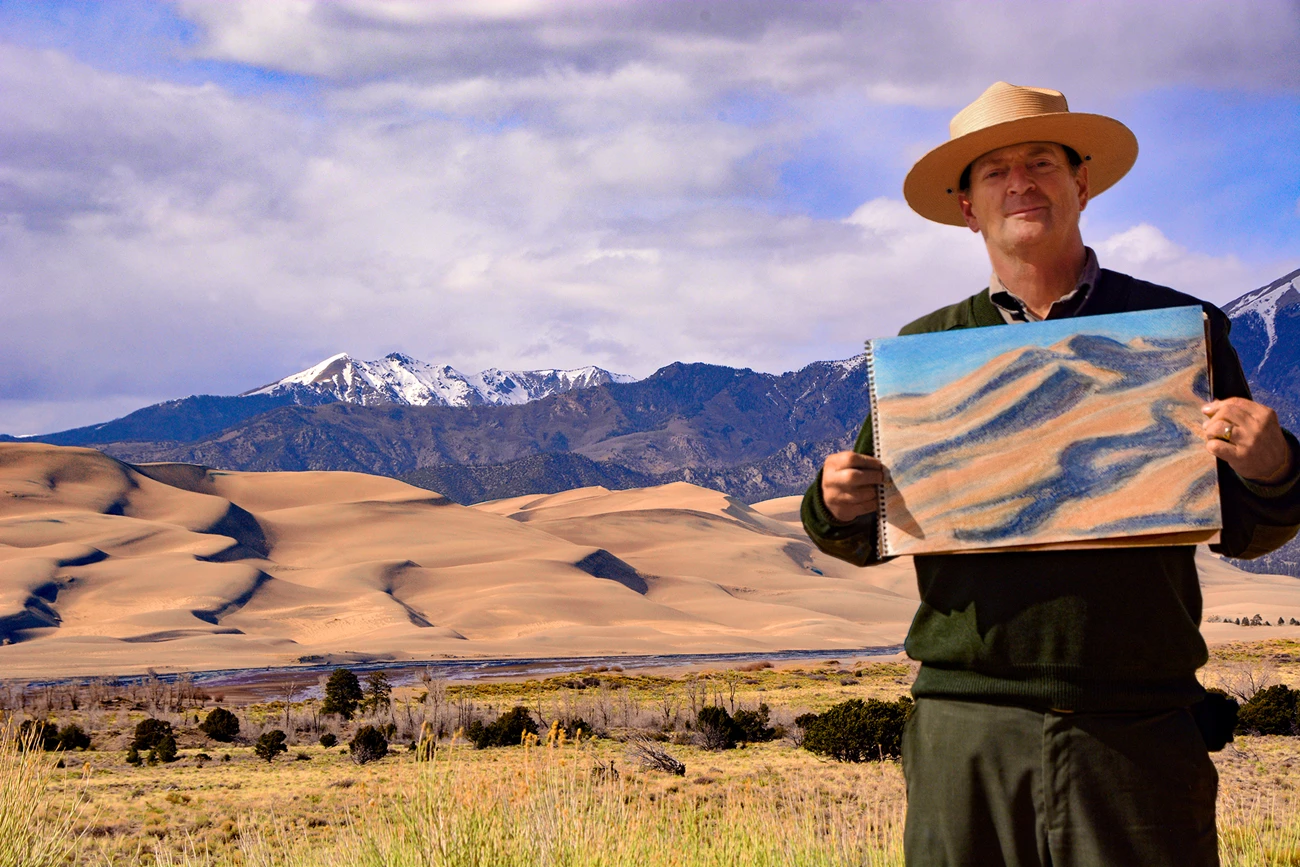Finished Sketch with Ranger Patrick Park ranger holding a color sketch of the dunes, with the dunes and snowcapped mountain in the background