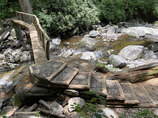 A set of wooden and gravel filled strais to a large foot loog bridge crossing a rushing stream.