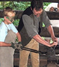 A student and ranger in the blacksmith shop. Both are using long metal tongs to mold the metal.