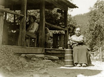 One of the sisters sits in front of a house, churning, and two other sisters are sitting on the porch