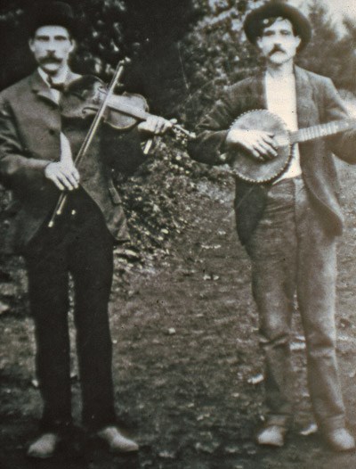 Historic photo of a man with a fiddle standing beside a man with a banjo.