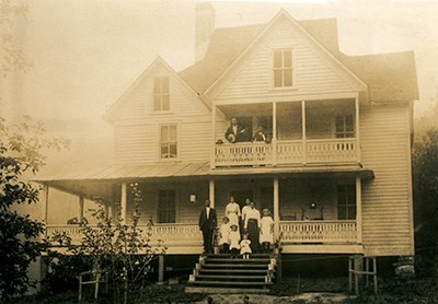 A large family stands on the upper and lower porches of their large Victorian home.