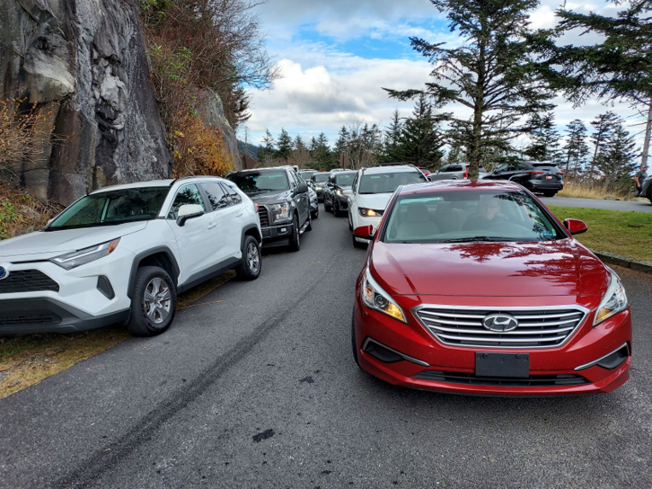 Vehicles parked illegally along the road shoulder and vehicles driving past in the lane of travel.