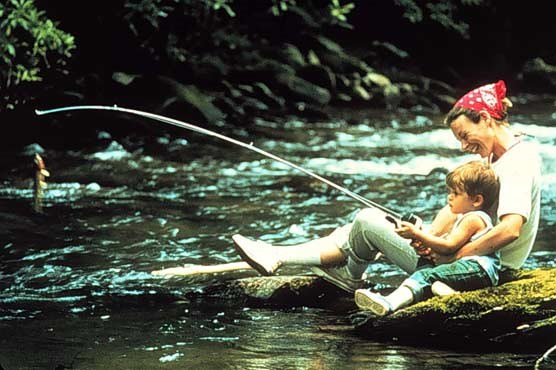 A photograph of a mother and son fishing a park stream.  A fish is hooked on the line of the fishing pole.