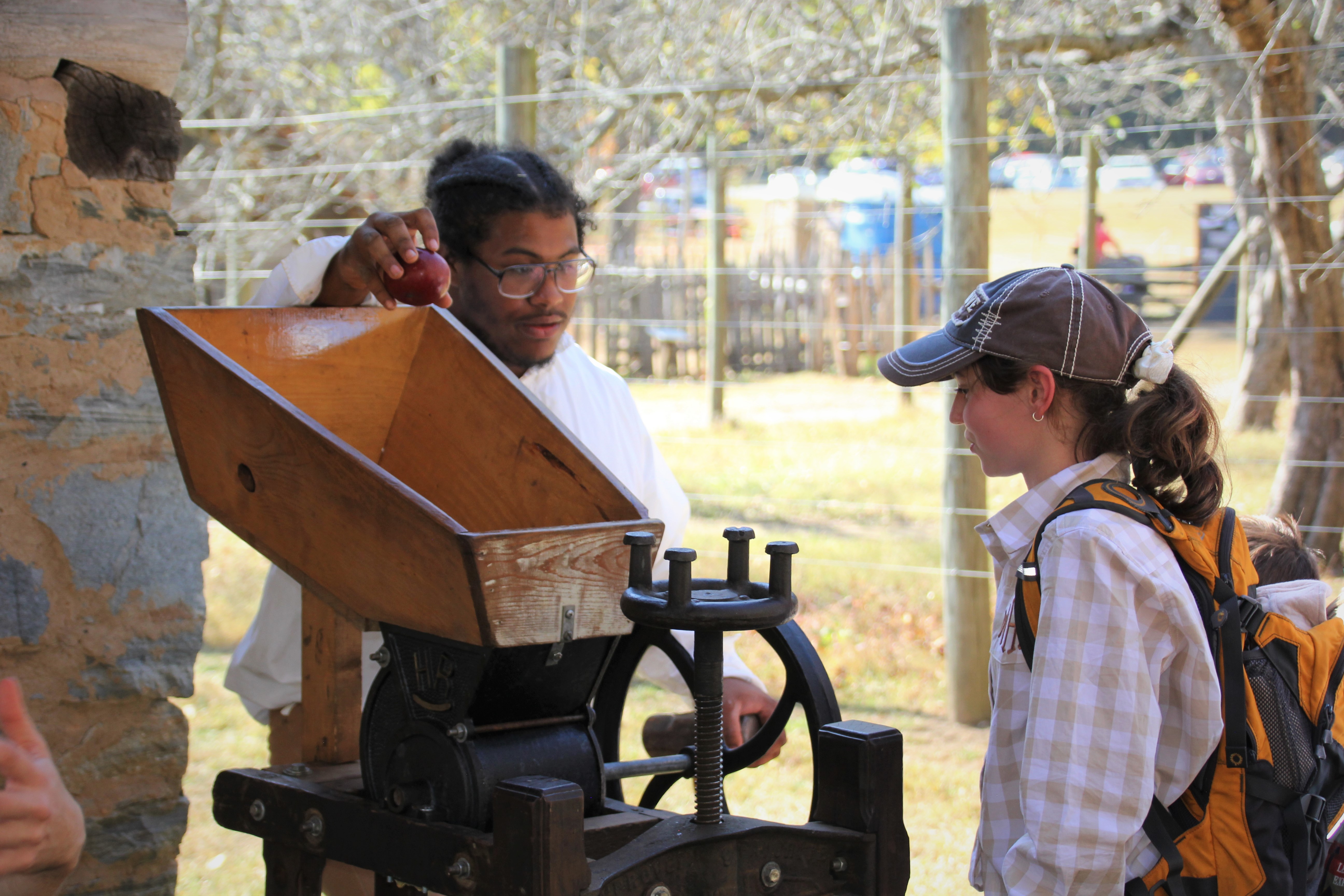 A person demonstrates an apple cider press to an onlooking child. The press has a wooden opening at the top for apples and metal components below.