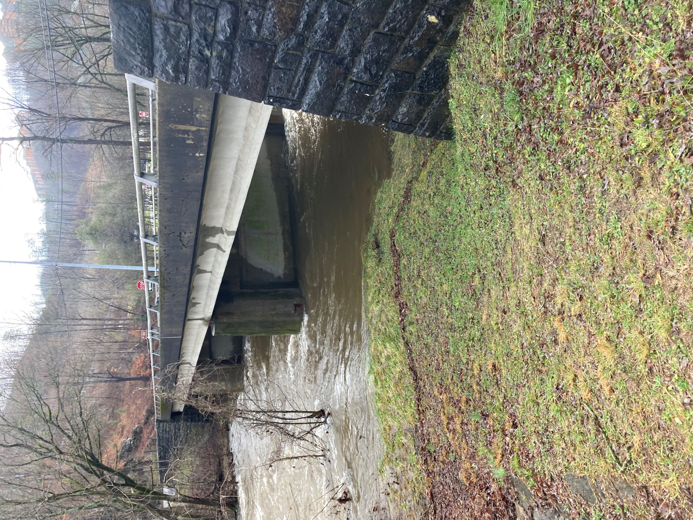11.15.22 Spur_Wiley Oakley Bridge_side view A side view of a bridge anchored with stone pillars and supports. A grassy bank lines the river flowing beneath.